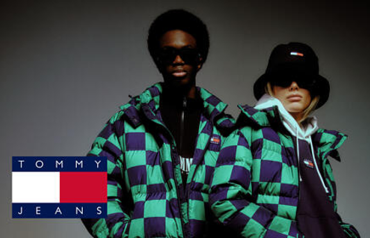 Zwei Models in grün-lila karierten Jacken, schwarzen Hüten und Sonnenbrillen stehen nebeneinander vor einem dunklen Hintergrund. Links ist das Tommy Jeans-Logo zu sehen.