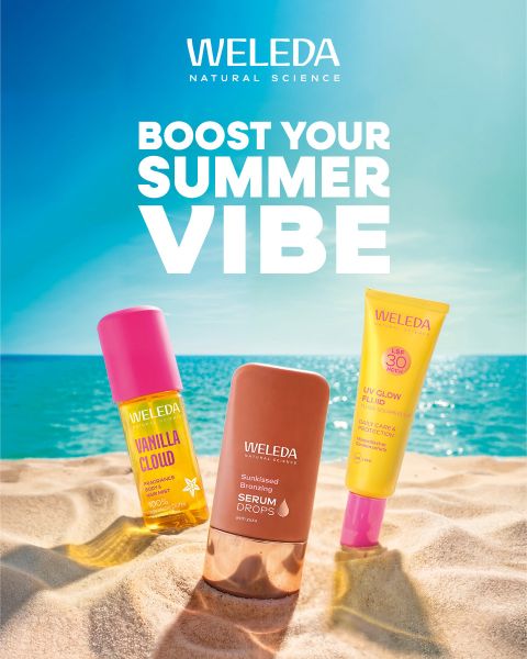 Drei Weleda Hautpflegeprodukte im Sand vor dem Hintergrund von Meer und blauem Himmel. Text: "Boost Your Summer Vibe". Helle, sommerliche Strandszene.