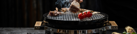 Auf einem Tischgrill mit Metallgitter werden Fleischstücke und ein Spieß mit Kirschtomaten gegrillt. Rauch steigt auf, während eine Hand das Essen mit einer Zange wendet. Gegrilltes Gemüse liegt auf einer Holzfläche in der Nähe.