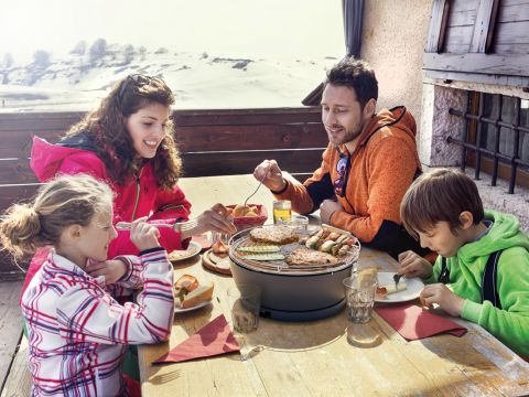 Eine vierköpfige Familie genießt ihr Essen auf einer sonnigen Terrasse, umgeben von einer verschneiten Landschaft. Sie sitzen warm angezogen um einen tragbaren Grill mit Essen herum. Die Atmosphäre ist fröhlich und entspannt.