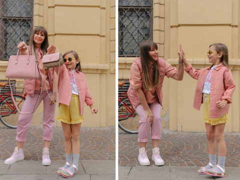 Eine Frau und ein junges Mädchen in aufeinander abgestimmten rosa und gelben Outfits teilen einen freudigen Moment. Im ersten Bild hält die Frau rosa Taschen, im zweiten geben sie sich ein High-Five. Im Hintergrund sind ein Fahrrad und ein Backsteingebäude zu sehen.