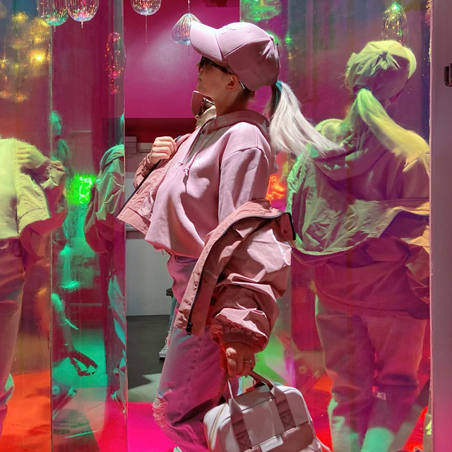 Eine Person in einem rosa Outfit und einer Mütze posiert in einem Raum mit bunten, reflektierenden Oberflächen. Sie hält eine Tasche und blickt nach oben. Das Licht wirft grüne, rosa und violette Reflexionen in den Raum.