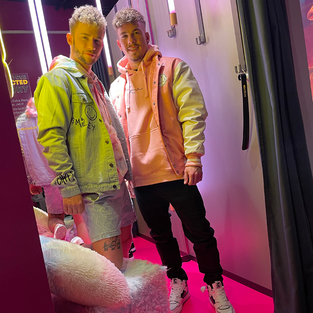 Zwei Männer posieren in einem hell erleuchteten Raum mit rosa Wänden. Einer trägt eine Jeansjacke und Shorts, der andere einen rosa Kapuzenpullover und Jeans. Sie stehen auf einem flauschigen rosa Teppich neben einer mit Lichtern geschmückten Wand.