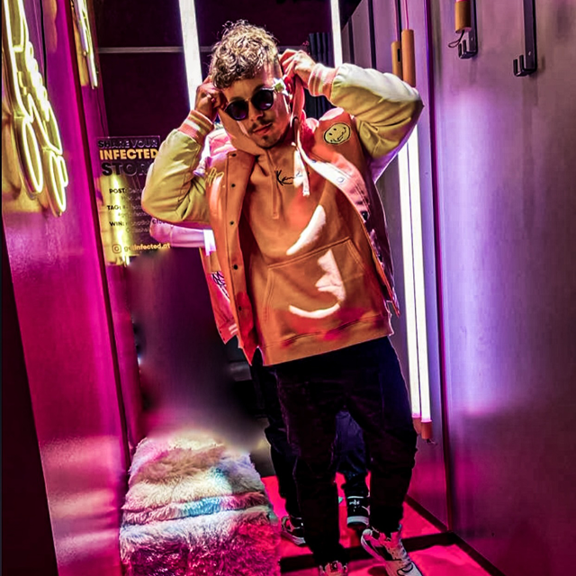 Eine Person mit Sonnenbrille, rosa Kapuzenpullover und Jacke steht in einem hell erleuchteten Raum mit Neonlicht. Der Raum ist farbenfroh eingerichtet, darunter ein pelziger Hocker und rosa Wände. Sie wirkt entspannt und stilvoll.