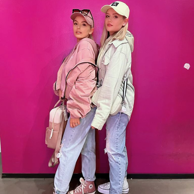 Zwei Menschen stehen vor einer leuchtend rosa Wand. Beide tragen lässige Outfits mit Baseballkappen, Sonnenbrillen und Rucksäcken. Sie posieren nach vorne gerichtet, einer in Rosatönen und der andere in Grau, mit Jeans und Turnschuhen.