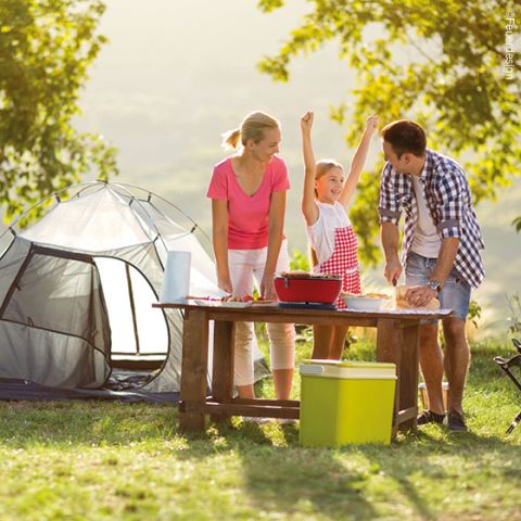 Eine Familie genießt einen sonnigen Tag auf einem Campingplatz. Im Hintergrund ist ein Zelt aufgebaut. Ein junges Mädchen mit erhobenen Armen steht zwischen zwei Erwachsenen an einem Tisch mit Campingausrüstung und Essen. Sie sind von Bäumen und Gras umgeben.