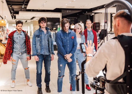Eine Gruppe von fünf Personen in Jeans-Outfits posiert gemeinsam in einem Geschäft. Ein Fotograf, ausgerüstet mit Kamera und Ausrüstung, macht ein Foto von ihnen. Die Umgebung ist hell und modern, im Hintergrund sind verschiedene Artikel ausgestellt.