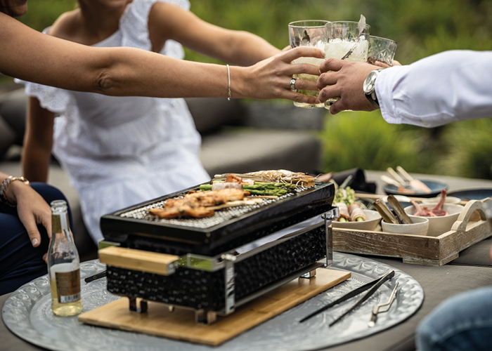 Menschen stoßen mit ihren Gläsern über einem Tischgrill an, auf dem Essen zubereitet wird, umgeben von Schüsseln und Besteck. Die Szene spielt im Freien mit viel Grün im Hintergrund und die Stimmung wirkt locker und gesellig.