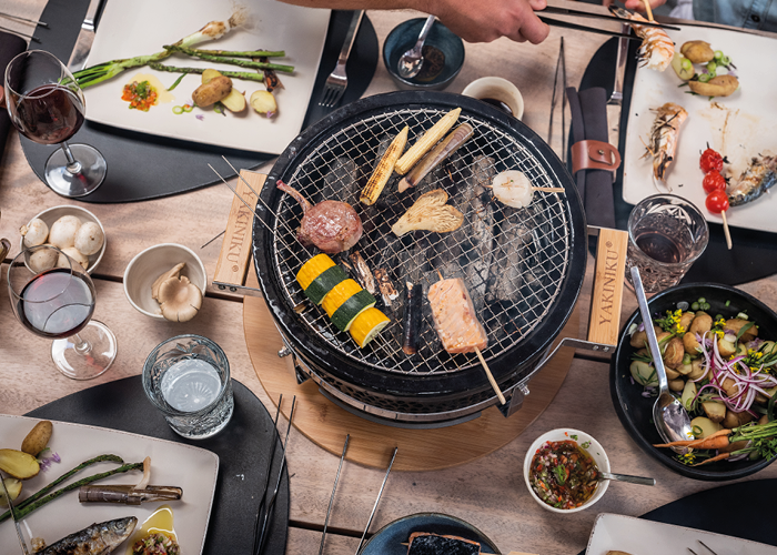 Eine Draufsicht auf einen Tischgrill mit verschiedenen Gemüsesorten und Fleischspießen. Um den Grill herum stehen Teller mit Grillgut, kleine Schüsseln mit Gewürzen, Gläser mit Wein und Wasser auf einem Holztisch. Mehrere Personen essen.