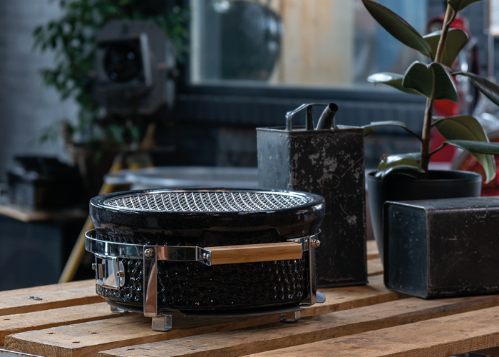 Auf einer Holzpalette steht ein runder, schwarzer tragbarer Grill mit Holzgriff. Daneben stehen ein schwarzer Metallbehälter und eine Topfpflanze mit breiten Blättern. Im Hintergrund ist ein Interieur im Industriestil mit einer unscharfen Kamera und einem Spiegel zu sehen.