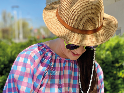 Eine Person mit Strohhut und Sonnenbrille steht draußen und blickt nach unten. Sie trägt ein buntes, kariertes Hemd. Im unscharfen Hintergrund sind grüne Sträucher und ein Gebäude zu sehen.