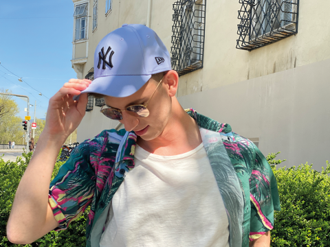 Eine Person mit Sonnenbrille, Tropenhemd und blauer Baseballkappe rückt ihren Hut zurecht, während sie an einem sonnigen Tag im Freien vor einem cremefarbenen Gebäude mit dekorativen Fenstergittern steht.