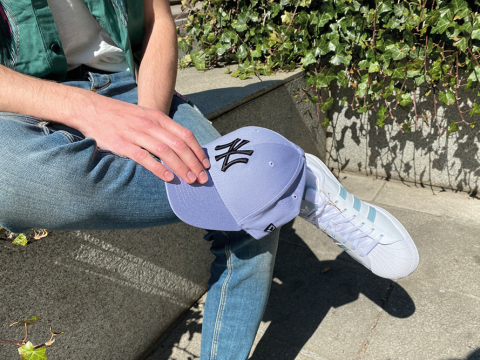 Eine Person sitzt auf einem Felsvorsprung. Sie trägt blaue Jeans, weiße Adidas-Sneaker mit hellblauen Streifen und hält eine pastellblaue New York Yankees-Kappe in der Hand. Im Hintergrund sind Efeublätter und Sonnenlicht zu sehen.