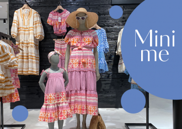 Eine Kleiderschau mit einer Schaufensterpuppe in einem bunten, gemusterten Kleid und Hut, mit einem passenden Kinderkleid auf einer anderen Schaufensterpuppe. Im Hintergrund sind weitere Kleider zu sehen. In einer Sprechblase steht „Mini me“ und blaue Kreise schmücken die Szene.