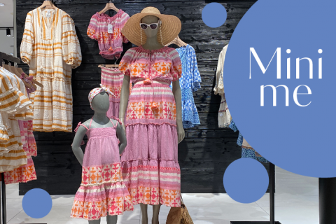 Eine Kleiderschau mit einer Schaufensterpuppe in einem bunten, gemusterten Kleid und Hut, mit einem passenden Kinderkleid auf einer anderen Schaufensterpuppe. Im Hintergrund sind weitere Kleider zu sehen. In einer Sprechblase steht „Mini me“ und blaue Kreise schmücken die Szene.