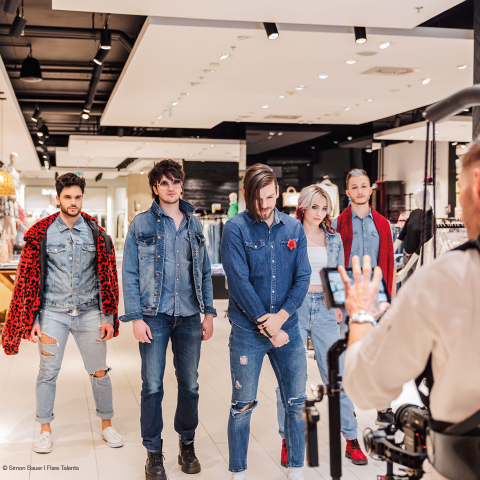 Eine Gruppe von fünf modisch gekleideten Menschen steht in einem Bekleidungsgeschäft. Sie tragen lässige Jeans-Outfits. Ein Fotograf macht ein Foto von ihnen. Das Geschäft ist mit Kleiderständern und heller Beleuchtung ausgestattet.