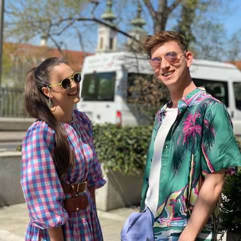 Eine Frau in einem karierten Kleid und Sonnenbrille lächelt einen Mann in einem Tropenhemd und einer Sonnenbrille an. Sie stehen an einem sonnigen Tag im Freien, im Hintergrund sind ein weißer Lieferwagen und eine von Bäumen gesäumte Straße zu sehen.