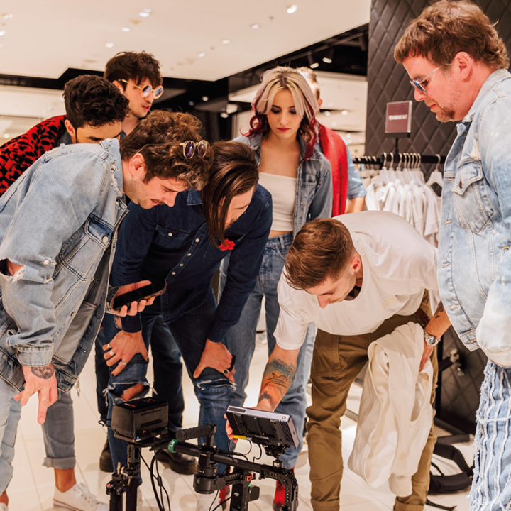 Eine Gruppe von acht Personen versammelt sich um eine Kamera in einem Geschäft. Sie sind leger gekleidet, manche tragen Jeans. Im Hintergrund sind Kleiderständer zu sehen.