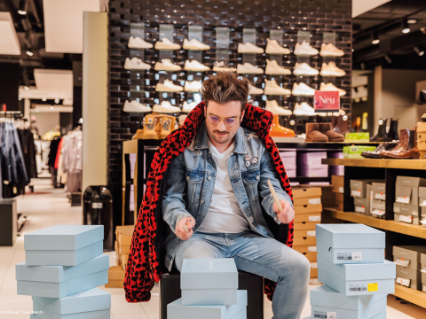Eine Person im Jeans-Outfit und einer rot-schwarzen Jacke sitzt auf einem Hocker in einem Schuhgeschäft, umgeben von blauen Schuhkartons. Im Hintergrund säumen Regale mit verschiedenen Schuhen die Wände.