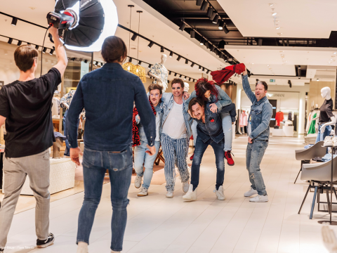 Eine Gruppe energiegeladener Menschen, einige in Jeans-Outfits, haben Spaß und posieren für ein Foto in einem hell erleuchteten Bekleidungsgeschäft. Eine Person wird Huckepack getragen. Ein Fotograf hält den lebhaften Moment fest.