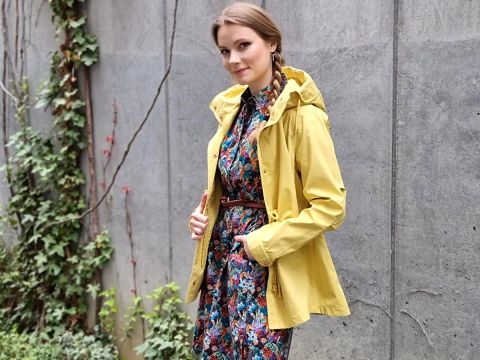 Eine Frau steht im Freien und trägt ein bunt gemustertes Kleid mit einem braunen Gürtel und einer gelben Jacke. Sie hat zwei Zöpfe und posiert mit den Händen auf ihrer Jacke. Im Hintergrund ist eine Betonwand mit Efeu und Grünpflanzen zu sehen.