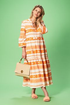 Eine Frau in einem langen, gemusterten Kleid mit orangen und weißen Mustern steht auf grünem Hintergrund. Sie hält eine beige gewebte Handtasche in einer Hand und trägt dazu passende Sandalen. Ihre Haare sind offen und sie blickt leicht zur Seite.