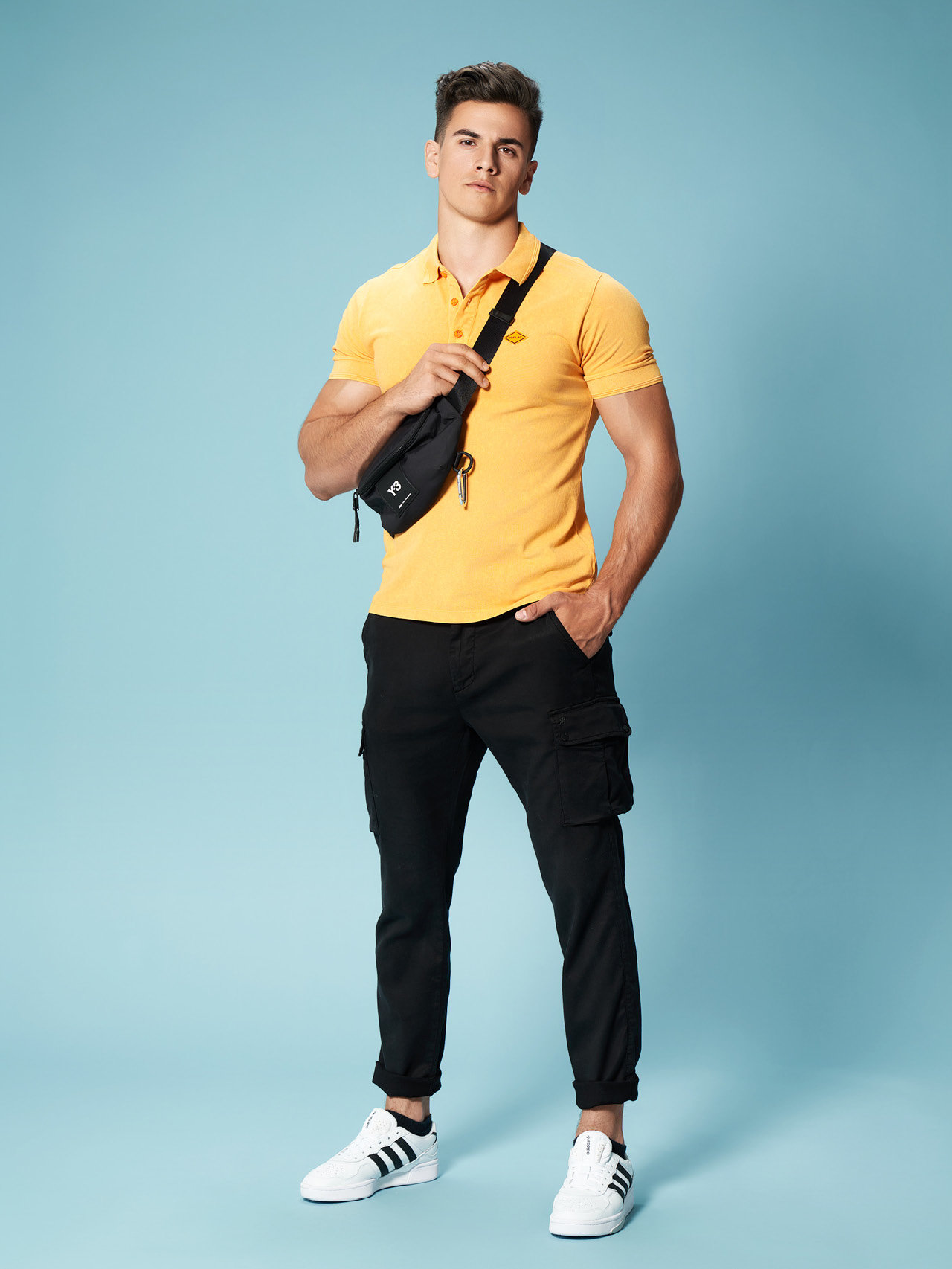 Ein Mann steht selbstbewusst vor einem hellblauen Hintergrund. Er trägt ein gelbes Poloshirt, schwarze Cargohosen und weiße Turnschuhe. Über seiner Schulter hängt eine schwarze Umhängetasche, und seine Hände stecken in den Taschen.