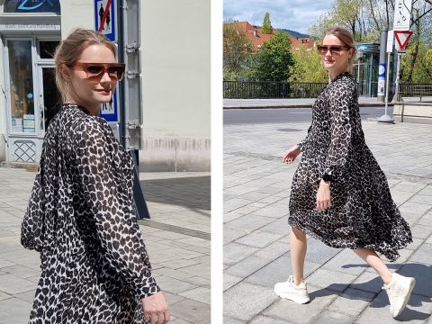 Eine Person mit Sonnenbrille und fließendem Kleid mit Leopardenmuster läuft auf einer sonnigen Straße. Sie trägt helle Turnschuhe und wurde in Bewegung aufgenommen. Im Hintergrund sind ein Gebäude und Grünflächen zu sehen.