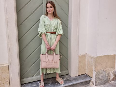 Eine Frau in einem hellgrünen Kleid steht vor einer grauen Tür. Sie hält eine hellrosa Einkaufstasche mit der Aufschrift „THE TOTE BAG“ und trägt hellbraune Sandalen. Im Hintergrund sind eine beige Wand und eine Marmorstufe zu sehen.