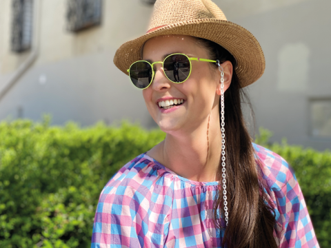 Frau mit Strohhut, grün umrandeter Sonnenbrille und bunt karierter Bluse lächelt, während sie im Freien sitzt. An ihrer Sonnenbrille baumelt eine Kette. Im Hintergrund sind grüne Büsche und eine Hauswand zu sehen.
