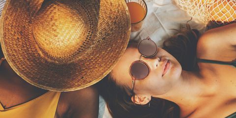 Zwei Menschen liegen am Strand. Einer trägt einen großen Strohhut, der andere eine runde Sonnenbrille. Sie entspannen auf einer weißen Decke und neben ihnen steht ein Glas Orangensaft.