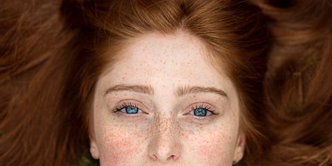 Nahaufnahme des Gesichts einer Person mit blauen Augen, Sommersprossen und ausgebreiteten langen roten Haaren. Die Person blickt direkt in die Kamera.