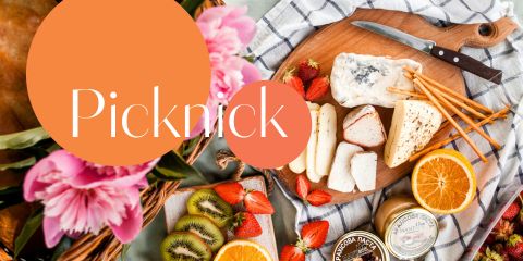 Ein Picknick-Set mit verschiedenen Käsesorten, einem Messer, Grissini, Erdbeeren, Orangenscheiben, Kiwi und blühenden Pflanzen. Über der Szene steht in großen Buchstaben das Wort „Picknick“.