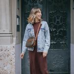Eine Frau steht vor einer verzierten Tür. Sie trägt eine Jeansjacke über einem braunen Kleid und trägt eine Umhängetasche. Sie hat schulterlanges Haar und lächelt, während sie zur Seite schaut.
