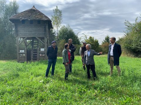 Eine Gruppe von Menschen steht auf einer Wiese vor einem Holzturm mit Strohdach. Die Szene spielt im Freien mit Bäumen im Hintergrund unter einem teilweise bewölkten Himmel.