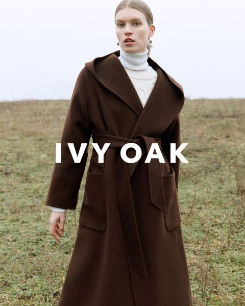 Eine Person, die einen langen braunen Mantel mit Gürtel trägt, steht auf einem offenen Feld. Sie trägt darunter einen weißen Rollkragenpullover und blickt nach vorne. Der Text „IVY OAK“ ist über das Bild gelegt. Der Hintergrund ist eine grasbewachsene, bewölkte Landschaft.
