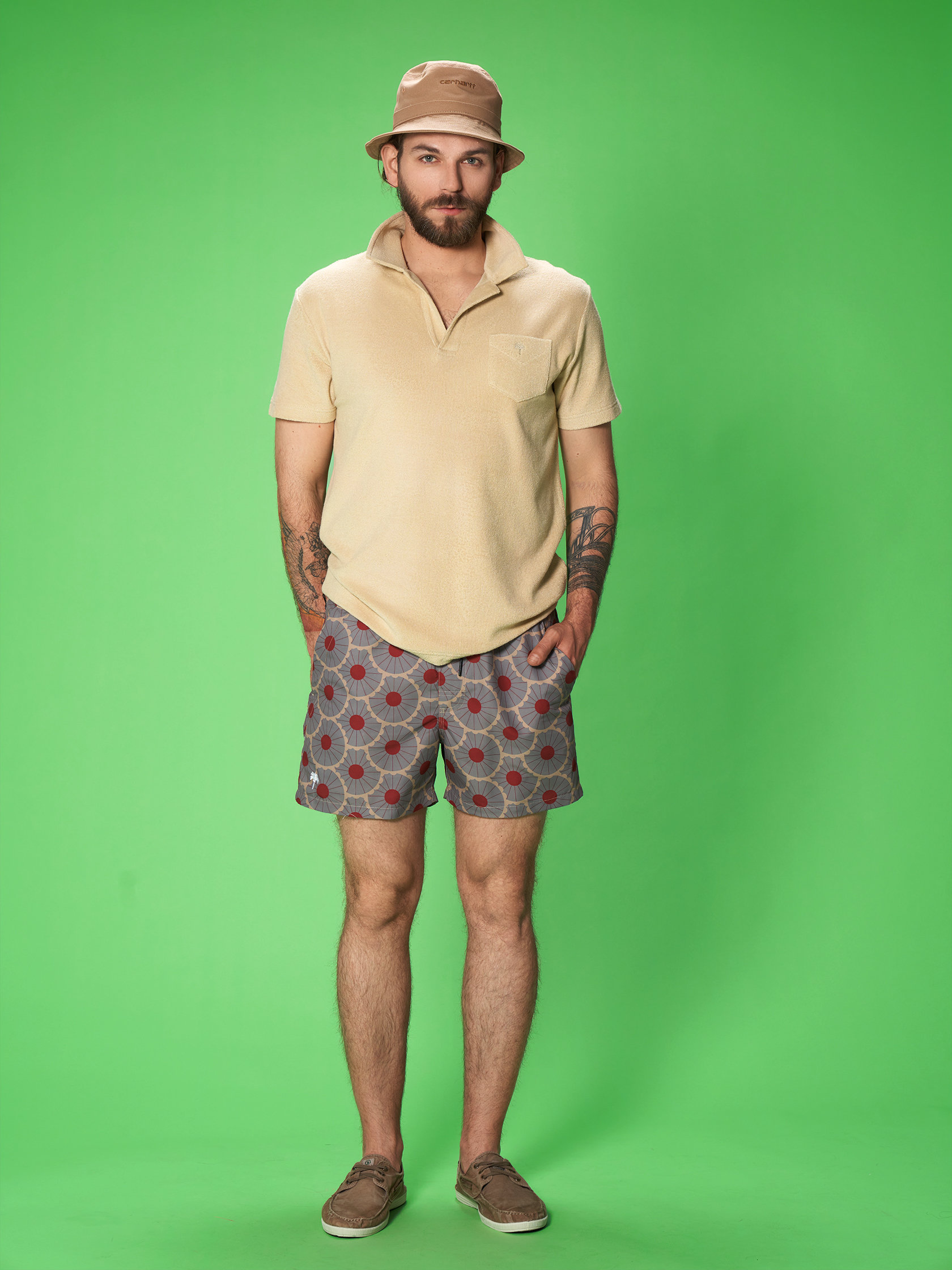 Ein Mann mit Bart und Tattoos steht vor einem grünen Hintergrund. Er trägt einen beigen Hut, ein beiges Kurzarmhemd und graue Shorts mit roten Punkten. Seine Hände stecken in den Taschen und er trägt braune Schuhe.