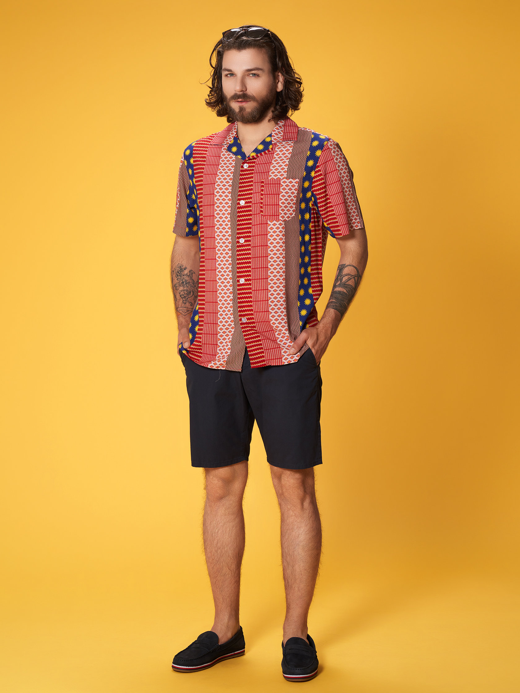 Ein Mann mit schulterlangem Haar steht vor einem gelben Hintergrund. Er trägt ein buntes, geometrisch gemustertes Kurzarmhemd, dunkle Shorts und schwarze Slipper. Seine Arme sind tätowiert und er hat einen Bart.