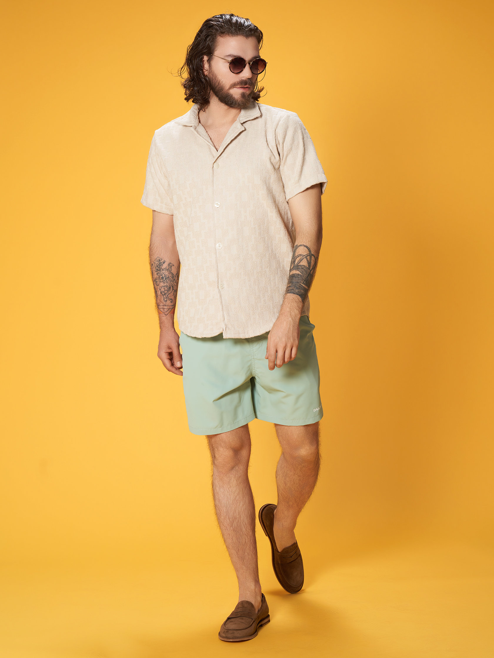 Eine Person mit langen Haaren und Sonnenbrille trägt ein beiges Kurzarmhemd, mintgrüne Shorts und braune Slipper. Sie hat Tattoos auf den Armen und steht vor einem orangefarbenen Hintergrund.