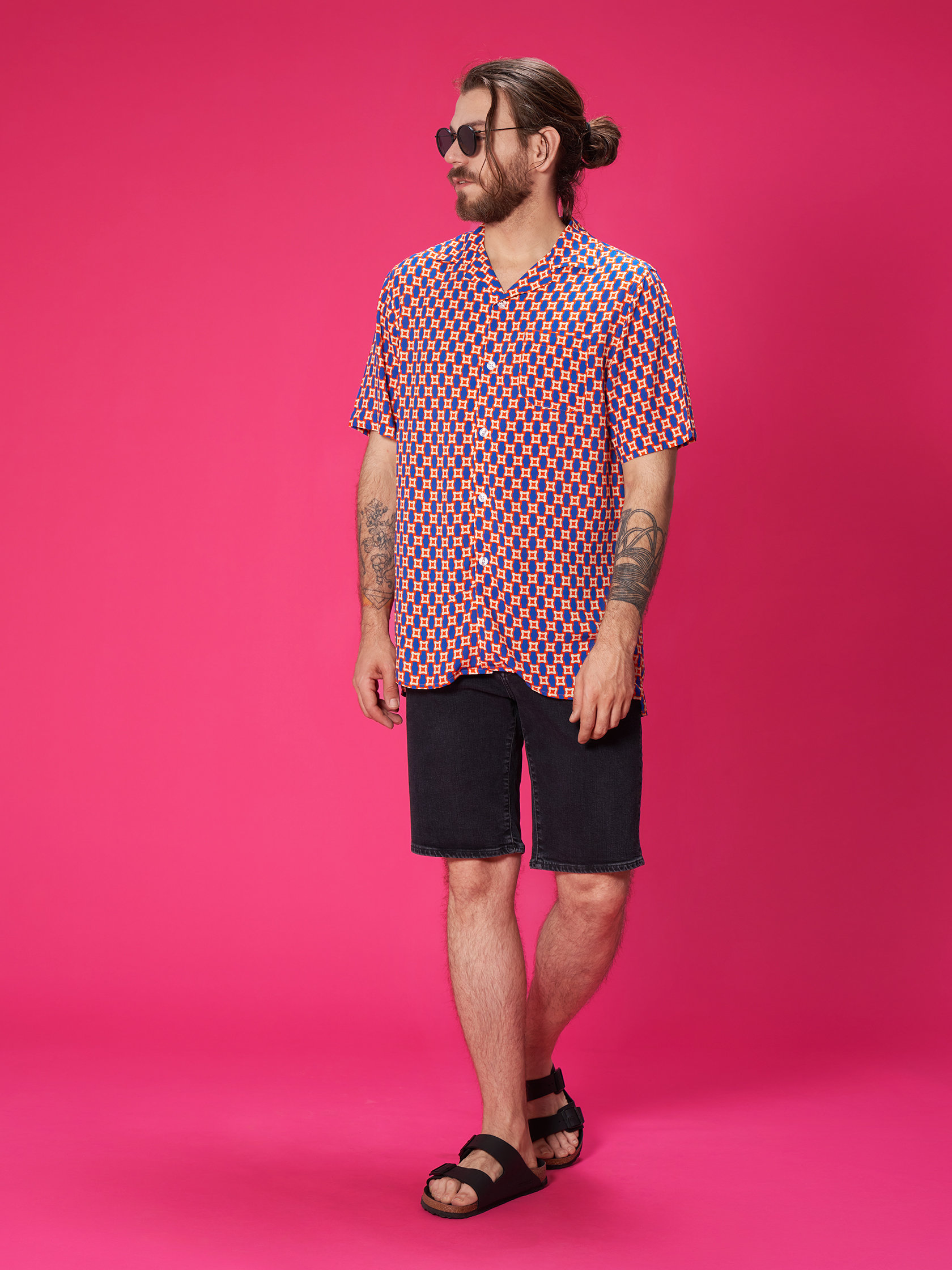 Ein Mann mit Dutt und Sonnenbrille steht vor einem rosa Hintergrund. Er trägt ein buntes Hemd mit geometrischem Muster, dunkle Shorts und schwarze Sandalen. Seine Arme sind tätowiert und er geht mit selbstbewusstem Schritt.