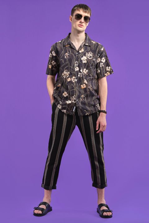 Eine Person steht vor einem violetten Hintergrund. Sie trägt eine Sonnenbrille, ein kurzärmeliges Hemd mit Blumenmuster, gestreifte Hosen und schwarze Sandalen. Ihre Hände stecken in den Taschen.