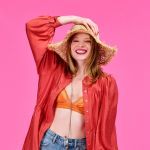 Eine Frau lächelt vor einem rosa Hintergrund. Sie trägt einen breitkrempigen Strohhut, ein orangefarbenes Oberteil und ein weites rotes Hemd. Sie hält den Hut mit einer Hand und trägt blaue Jeans.