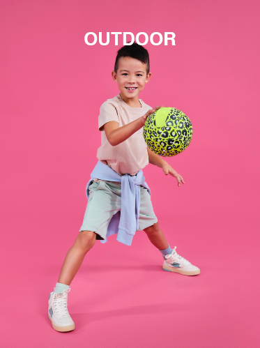 Ein kleiner Junge posiert voller Energie mit einem bunten Basketball vor einem leuchtend rosa Hintergrund. Er trägt ein hellrosa Hemd, hellblaue Shorts und weiße Turnschuhe. Über ihm prangt das Wort „OUTDOOR“.