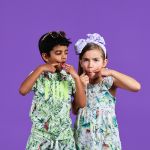 Zwei Kinder, die lustige Grimassen schneiden, beide tragen tropische Outfits. Der Junge links trägt eine Sonnenbrille und das Mädchen rechts ein großes Stirnband mit Schleife. Sie stehen vor einem einfarbig violetten Hintergrund.