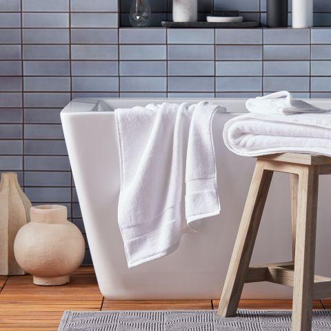 Modernes Badezimmer mit einer weißen Badewanne, die mit ordentlich gefalteten weißen Handtüchern geschmückt ist. Daneben steht ein Holzhocker mit weiteren Handtüchern. Im Hintergrund sind blaugrau geflieste Wände und der Boden ist aus Holz mit einem grauen Teppich.