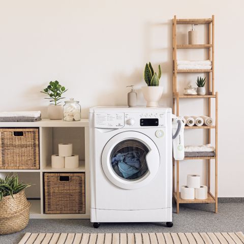 Eine aufgeräumte Waschküche mit einer weißen Frontlader-Waschmaschine und Wäsche darin. Um sie herum stehen geflochtene Körbe, Pflanzen und Regale mit ordentlich gestapelten Handtüchern und Toilettenpapierrollen. Auf dem Boden liegt ein gestreifter Teppich.