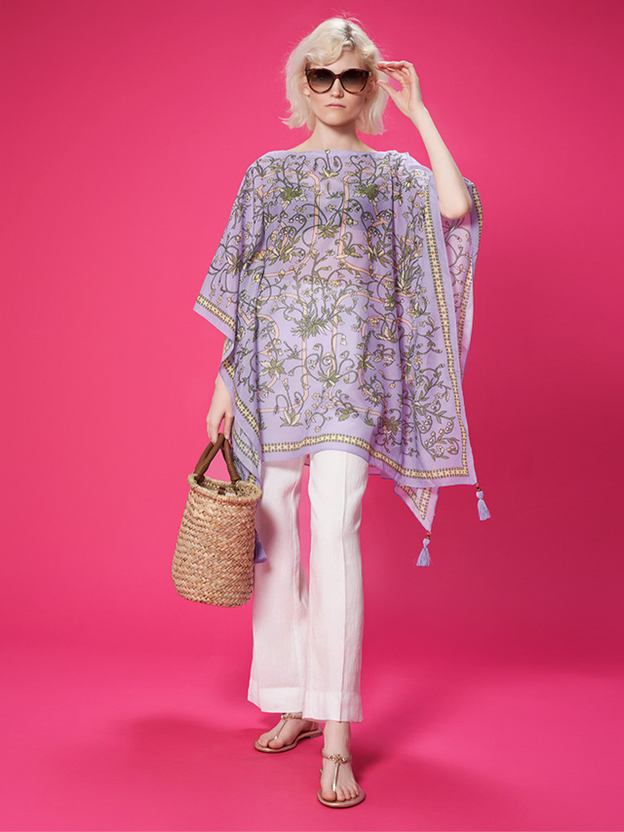 Eine Person mit Sonnenbrille, einem fließenden Kaftan mit lila Muster, weißen Hosen und Sandalen steht vor einem rosa Hintergrund. In einer Hand hält sie eine geflochtene Strohtasche.