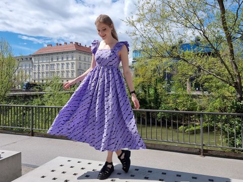 Eine Frau in einem lila Kleid mit Wellenmuster wirbelt auf einem Dach herum und bietet eine malerische Aussicht. Im Hintergrund sind Bäume und Gebäude unter einem teilweise bewölkten Himmel zu sehen. Sie lächelt und trägt schwarze Sandalen.