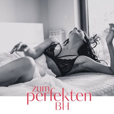 Eine Frau in einem Spitzen-BH liegt auf dem Bett, lächelt und hat den Kopf nach oben geneigt. Der Raum ist sanft beleuchtet. Der Text „zum perfekten BH“ ist über das Bild gelegt.