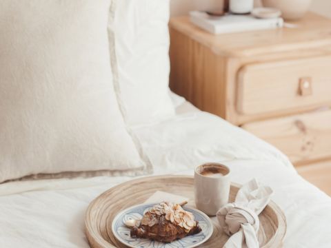 Eine gemütliche Schlafzimmerszene mit einem weißen Bett und einem Nachttisch aus Holz. Auf dem Bett steht auf einem Holztablett ein Teller mit einem mit Mandeln belegten Croissant, eine Tasse Kaffee und eine beige Serviette. Auf dem Tisch stehen ein Stapel Bücher und eine Kerze.